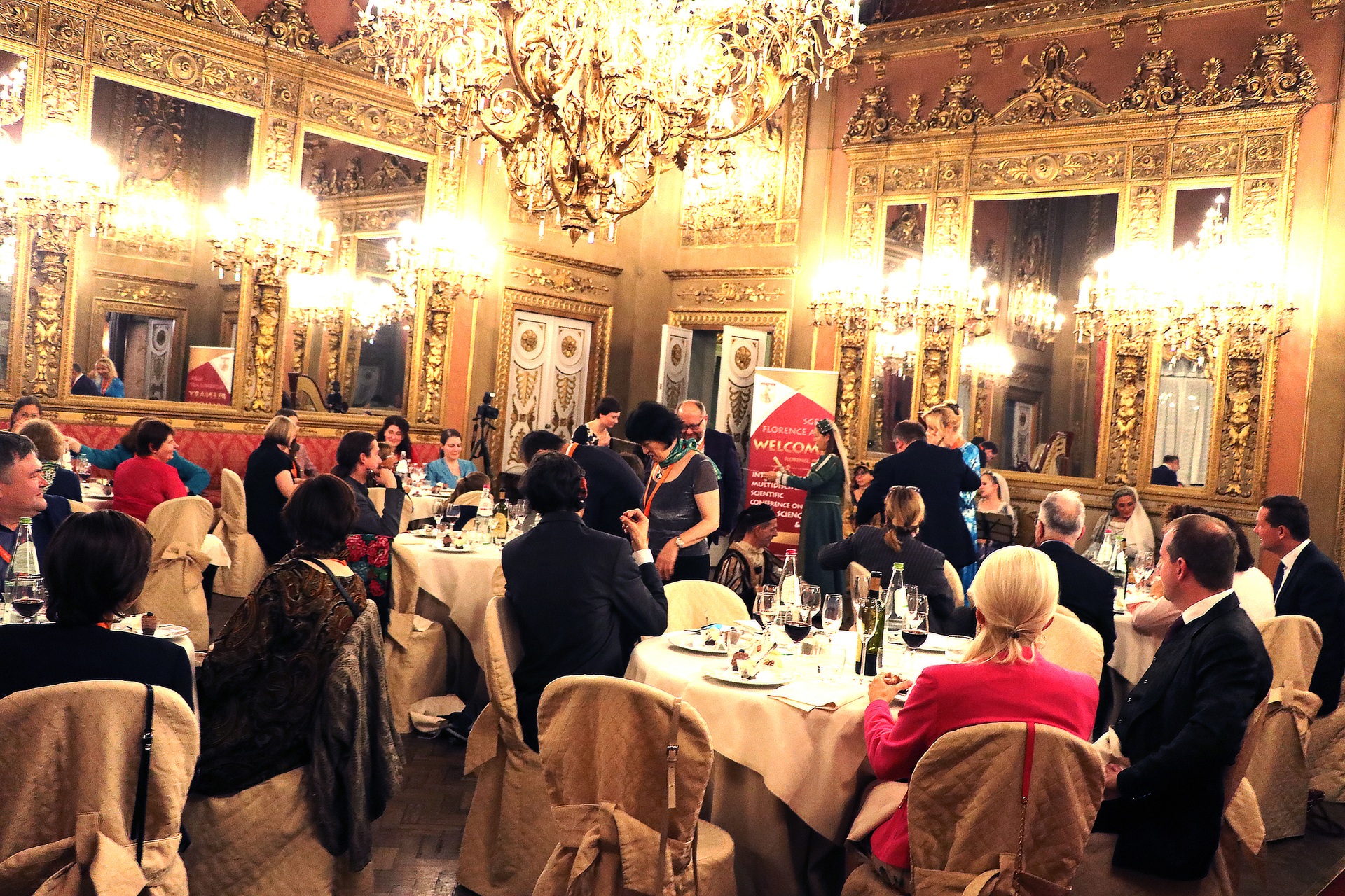 Invitados de la conferencia cenando en un salón dorado bajo candelabros